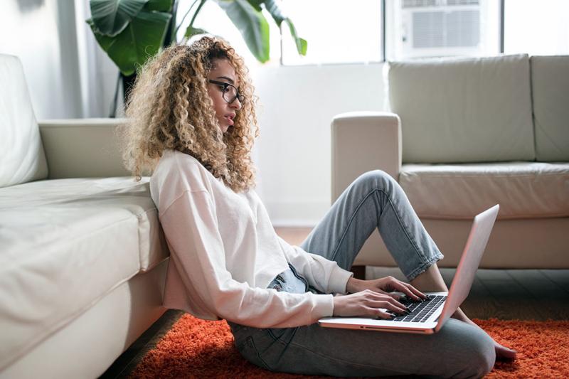 A student at home sits on the floor holding a laptop.