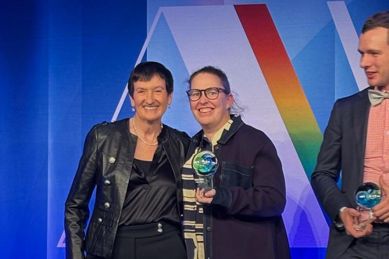 VU’s Tessa Benson is standing on a stage, lit up in lights holding a trophy, smiling, next to award presenter; Jennifer Westacott. Both are wearing black, with Tessa wearing a white striped shirt and denim jacket, with glasses.