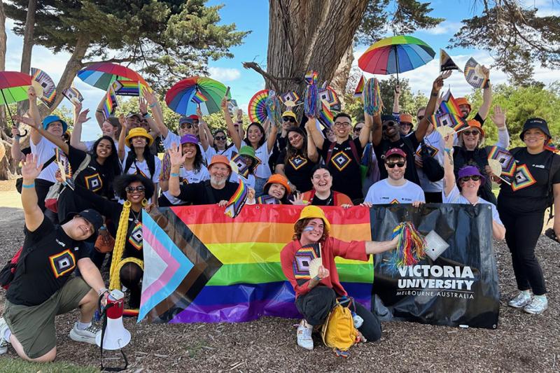 A group of smiling people in an Australian park with rainbow umbrellas, pride flags and Victoria University banners