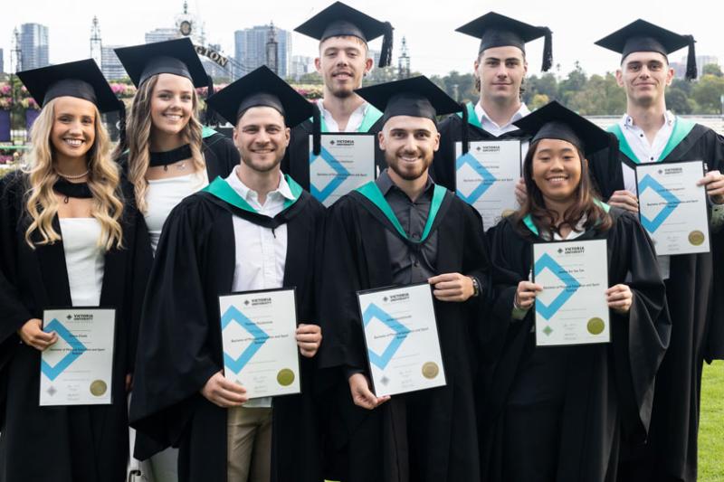 A group of 8 graduates smile proudly after their graduation ceremony at Flemington Racecourse