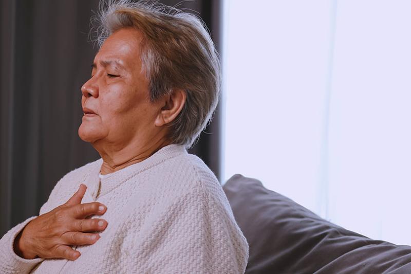 A mature-aged woman breathes in, she holds her hand to her chest, and her eyes are closed, indicating she is breathing with difficulty or experiencing pain