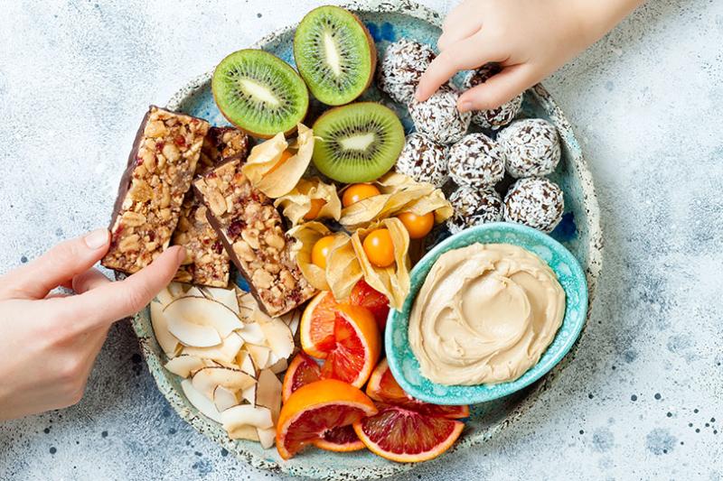 Two hands reach in to take food from a snack plate with granola bars, homemade raw energy balls, cashew butter, toasted coconut chips, fruits