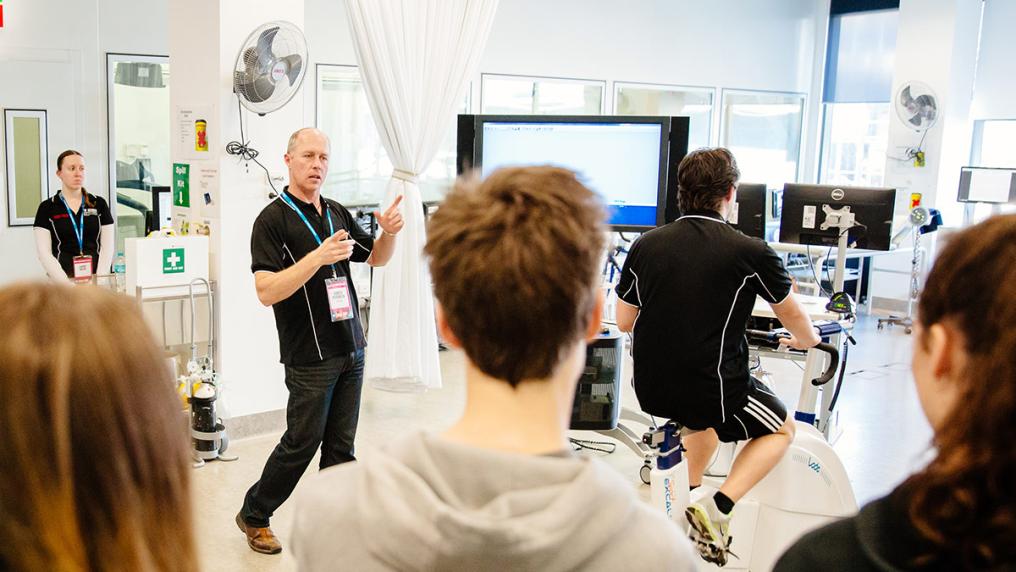 students watching a teacher as he explains as someone demonstrates on  an exercise bike