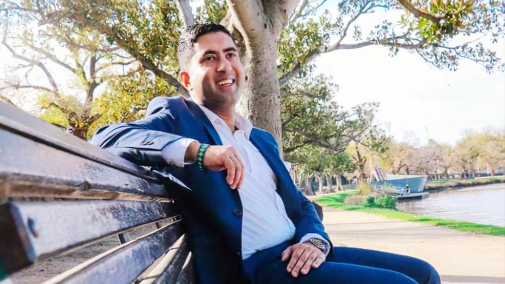 A man in a suit and shirt poses outdoors on a park bench