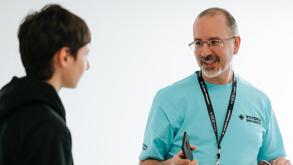 A middle aged man in a Victoria University t-shirt smiles and talks to a young person