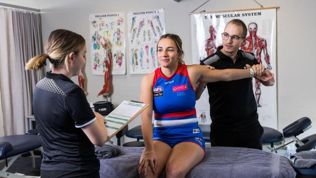 A person in a Western Bulldogs uniform having their arm stretched by a VU student. Another person is holding a clipboard and reading it.