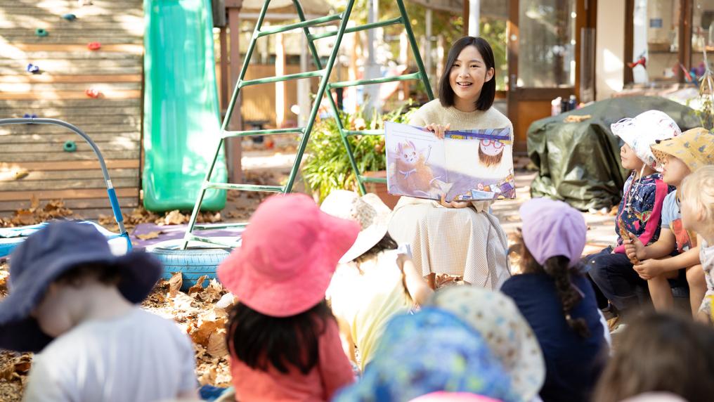 Early childhood educator reading a book to a class of pre-school children in the playground