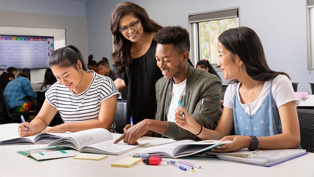 Three VU students sit working in a classroom, while being assisted by a teacher.