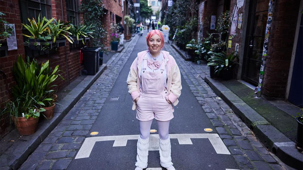A student wearing pink, with pink hair stands in the middle of a laneway in Melbourne city.