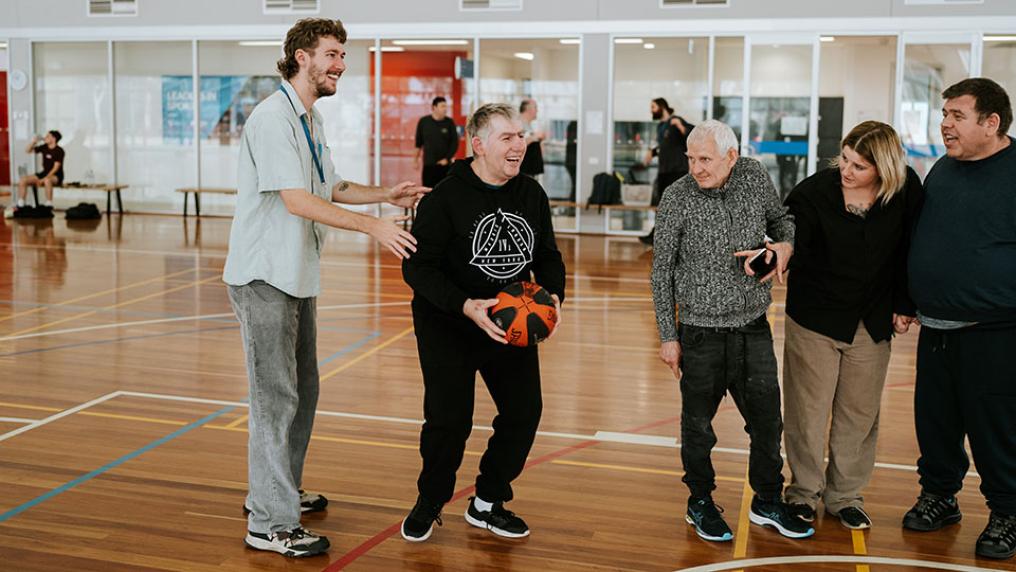A man holding a basketball preparing to shoot. A man stands behind him, holding him. Three people are watching by the man's side.
